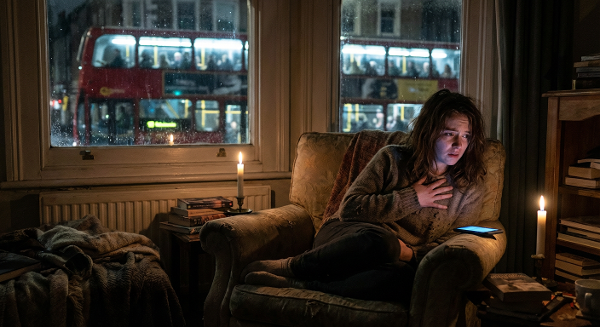 Девушка вечером в одиночестве испытывает тревогу, глядя на оживлённую улицу за окном.